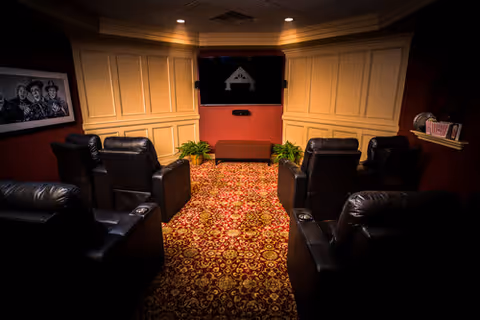 A cozy home theater room with six black leather recliners arranged in two rows facing a wall-mounted flat screen TV. The room has warm lighting, red and gold patterned carpet, cream-colored paneled walls, and a framed black and white photo on the left wall. Two green plants flank a small brown bench beneath the TV.