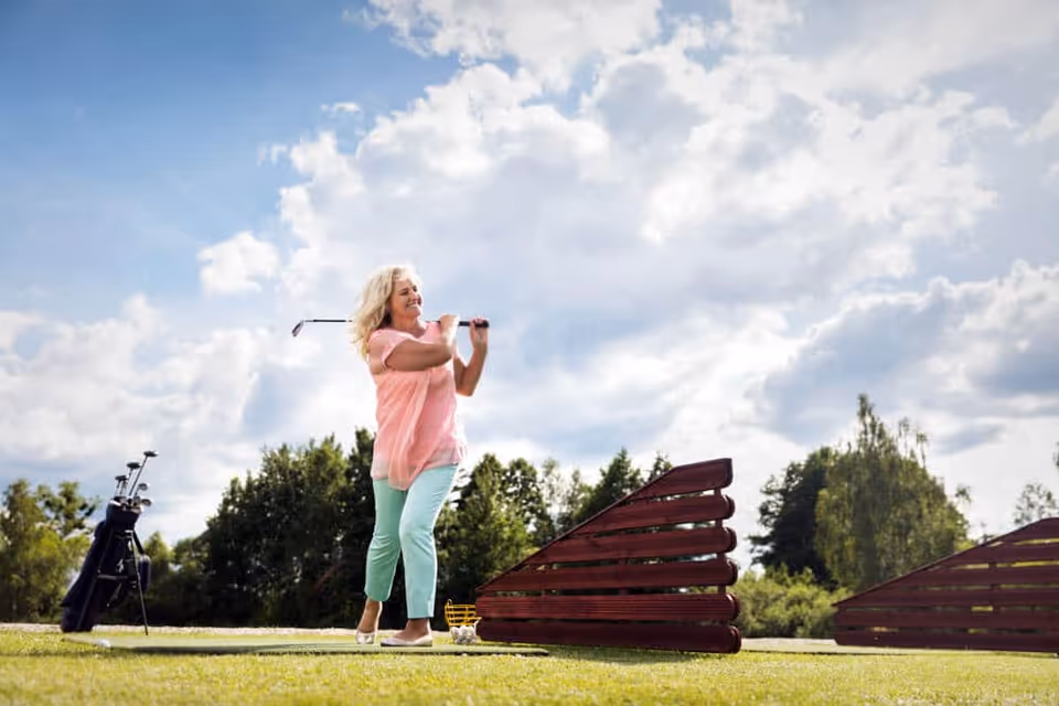 A woman in a pink top and light blue pants is playing golf outdoors on a sunny day with a partly cloudy sky. She is mid-swing with a golf club, standing on green grass with a golf bag and clubs nearby. Trees and wooden barriers are visible in the background.