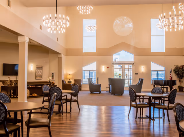 Spacious and well-lit common area in Carefield Pleasanton Memory Care featuring high ceilings with modern chandeliers, multiple round tables with chairs, a seating area with armchairs near large windows, a piano, and decorative plants.