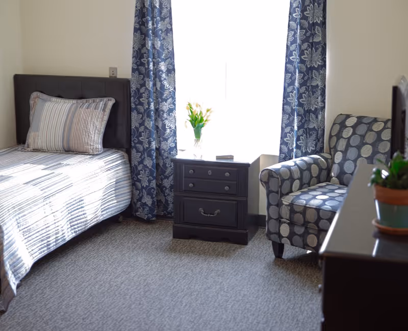 A bright bedroom with a single bed featuring a striped bedspread and a large pillow. Next to the bed is a dark wooden nightstand with drawers, holding a vase of flowers. Blue curtains with a floral pattern frame a window behind the nightstand. To the right, there is a cushioned armchair with a circular pattern, and part of a dark wooden dresser with a small potted plant is visible.