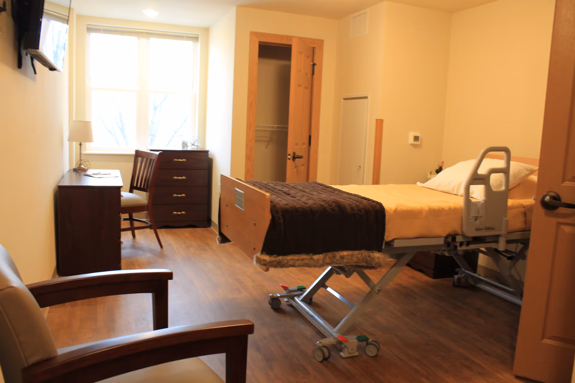 A well-lit senior living bedroom with a hospital-style adjustable bed covered with yellow bedding and a dark brown blanket. The room features wooden flooring, a wooden chair with armrests, a small desk with a chair, a chest of drawers near a window, and an open closet door.