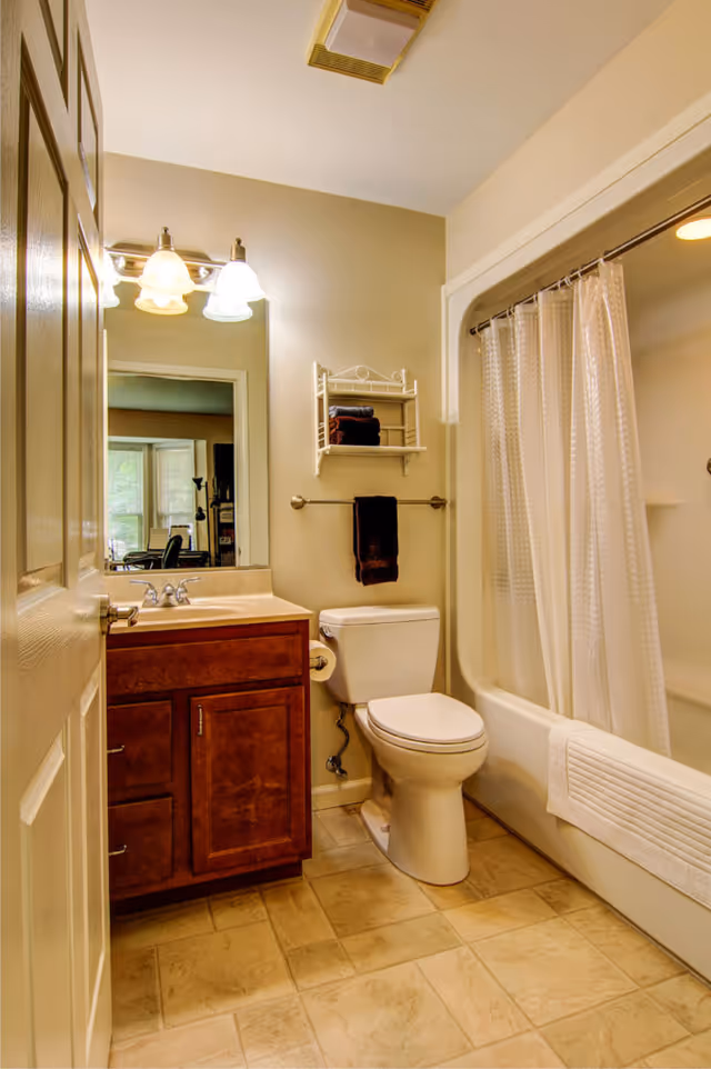 A clean bathroom with a wooden vanity cabinet, a sink with a mirror above it, a toilet, and a bathtub with a white shower curtain. There are towels hanging on a rack above the toilet and on the side of the bathtub. The floor is tiled and the walls are painted beige.