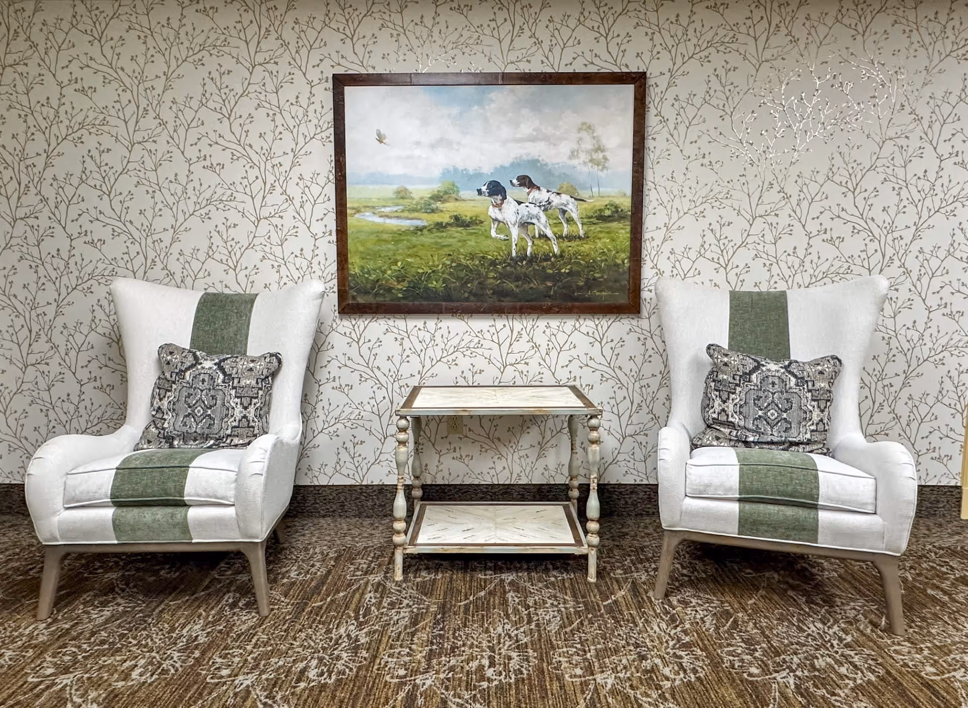 Two white armchairs with green vertical stripes and patterned cushions are positioned on either side of a small white wooden side table. Behind them is a wall with beige wallpaper featuring a subtle branch pattern, and a framed painting of two hunting dogs in a green field hangs above the table.