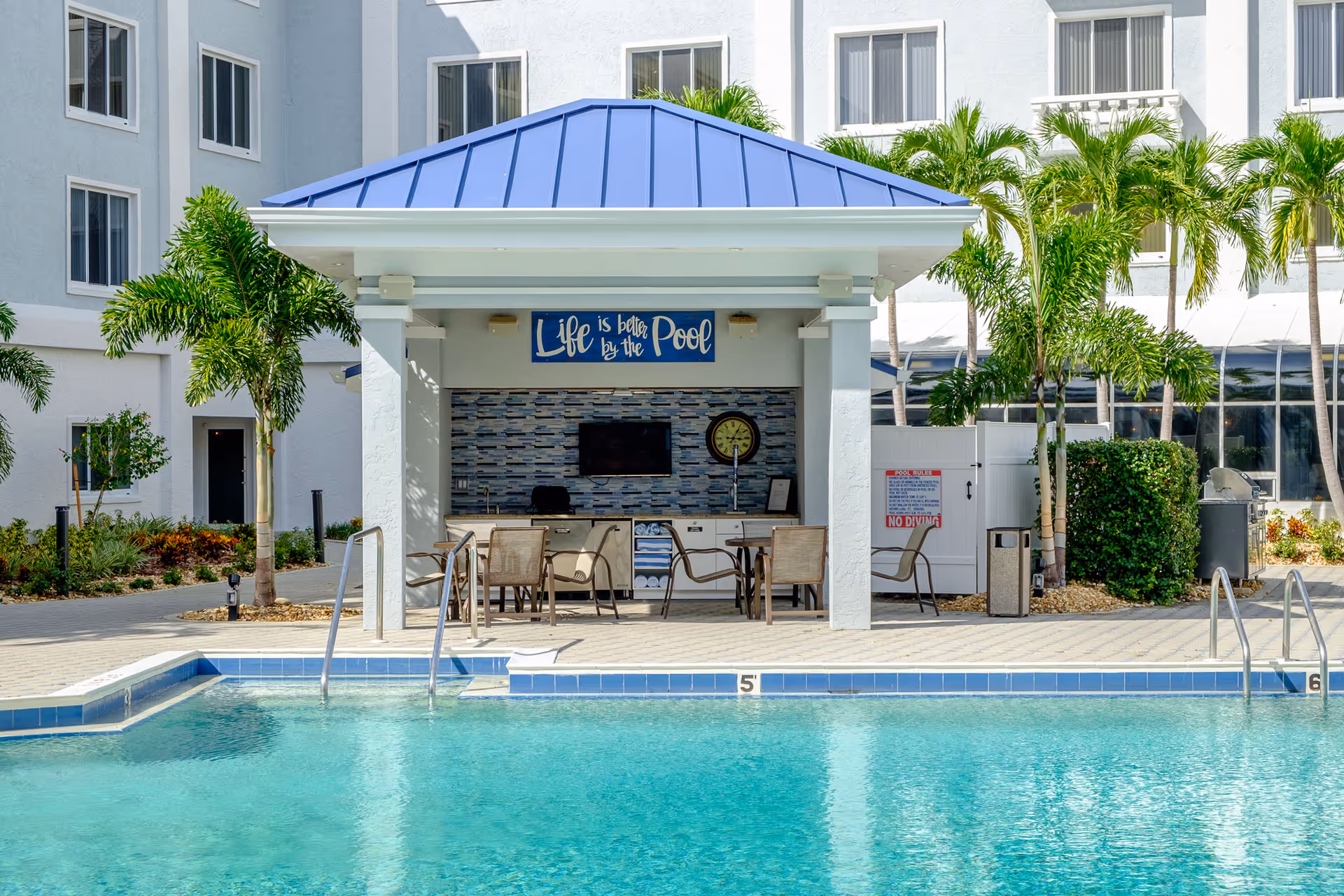 Outdoor swimming pool with a blue-roofed poolside pavilion reading 'Life is better by the Pool', chairs, and palm trees in front of a white building.