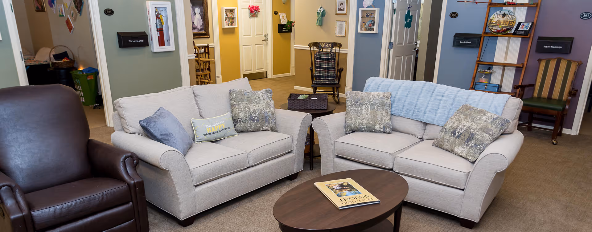 A senior living common area with two light-gray loveseats, a brown recliner, a coffee table with a book, decorative pillows and a hallway with doors and nameplates in the background.