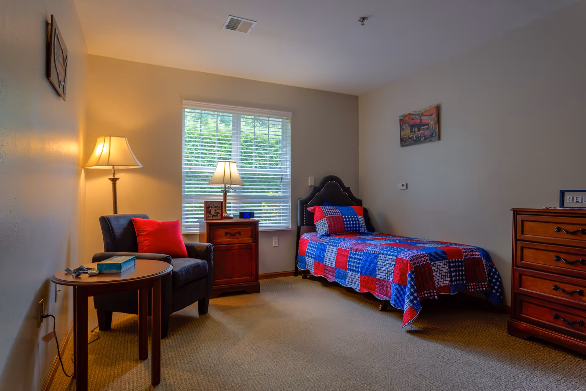 A cozy bedroom with a single bed covered in a red, blue, and white patchwork quilt. Next to the bed is a wooden nightstand with a lamp and a small framed sign. A comfortable armchair with a red pillow sits beside a round wooden table with a book and glasses on it. The room has beige walls, a window with white blinds, and a wooden dresser with drawers. Two lamps provide warm lighting.