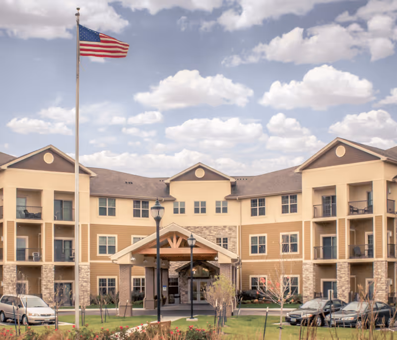 Exterior view of a three-story senior living facility building with balconies, an entrance canopy, parked cars, a flagpole with an American flag, and a partly cloudy sky.