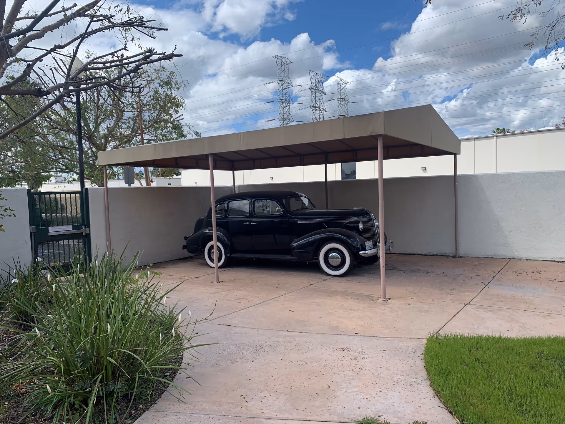 A vintage black car with whitewall tires parked under a beige metal carport in an outdoor area with concrete pavement, green grass, and some plants. There is a white wall surrounding the area and a partly cloudy sky with power lines and towers in the background.