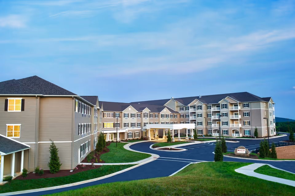 Exterior view of a large, multi-story senior living facility building with beige siding and multiple windows. The building is surrounded by landscaped grass, small trees, and a paved driveway with parking spaces. The sky is clear with a soft blue hue.