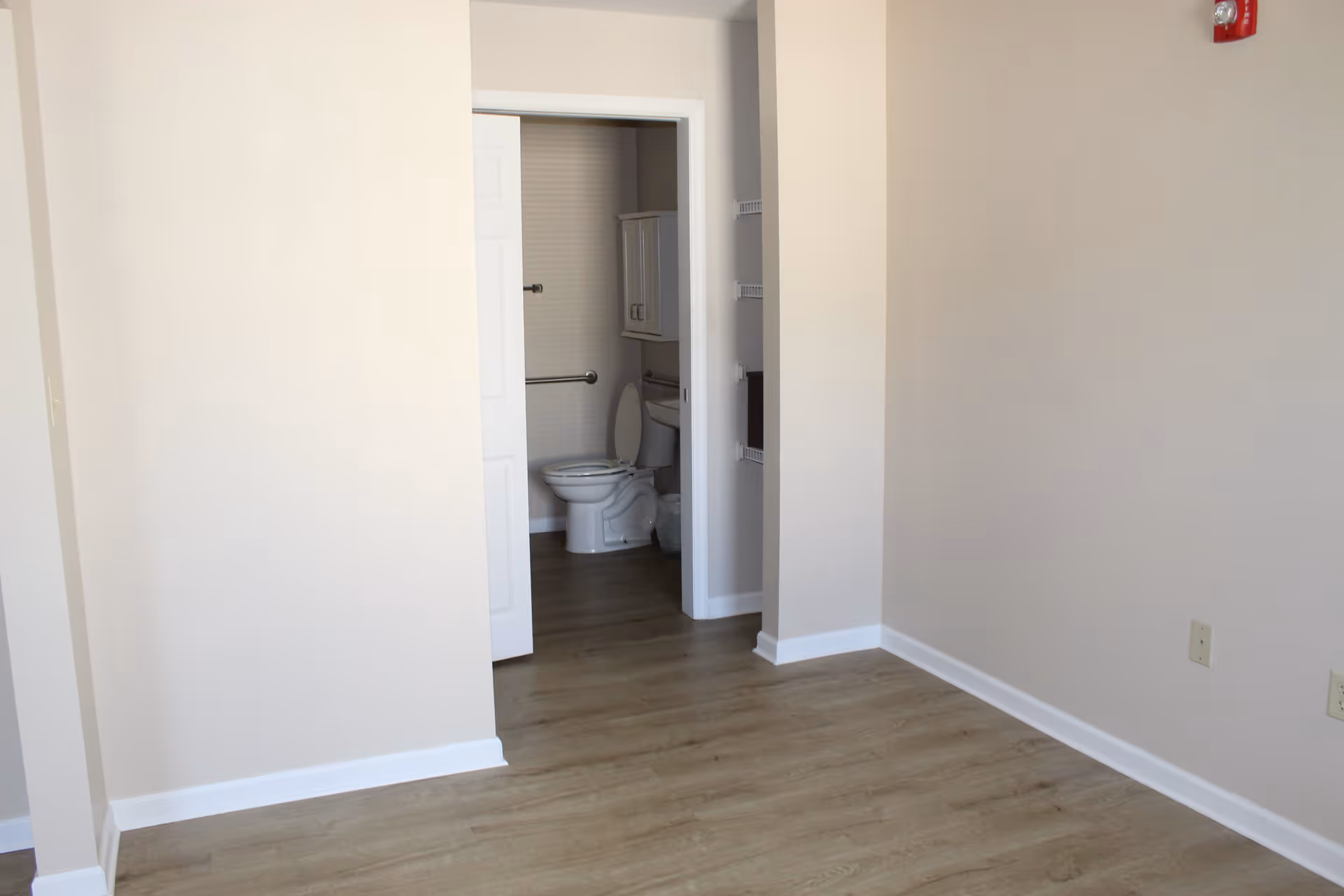 Empty room with light-colored walls and wood flooring, showing an open doorway leading to a bathroom with a visible toilet and wall-mounted cabinet.