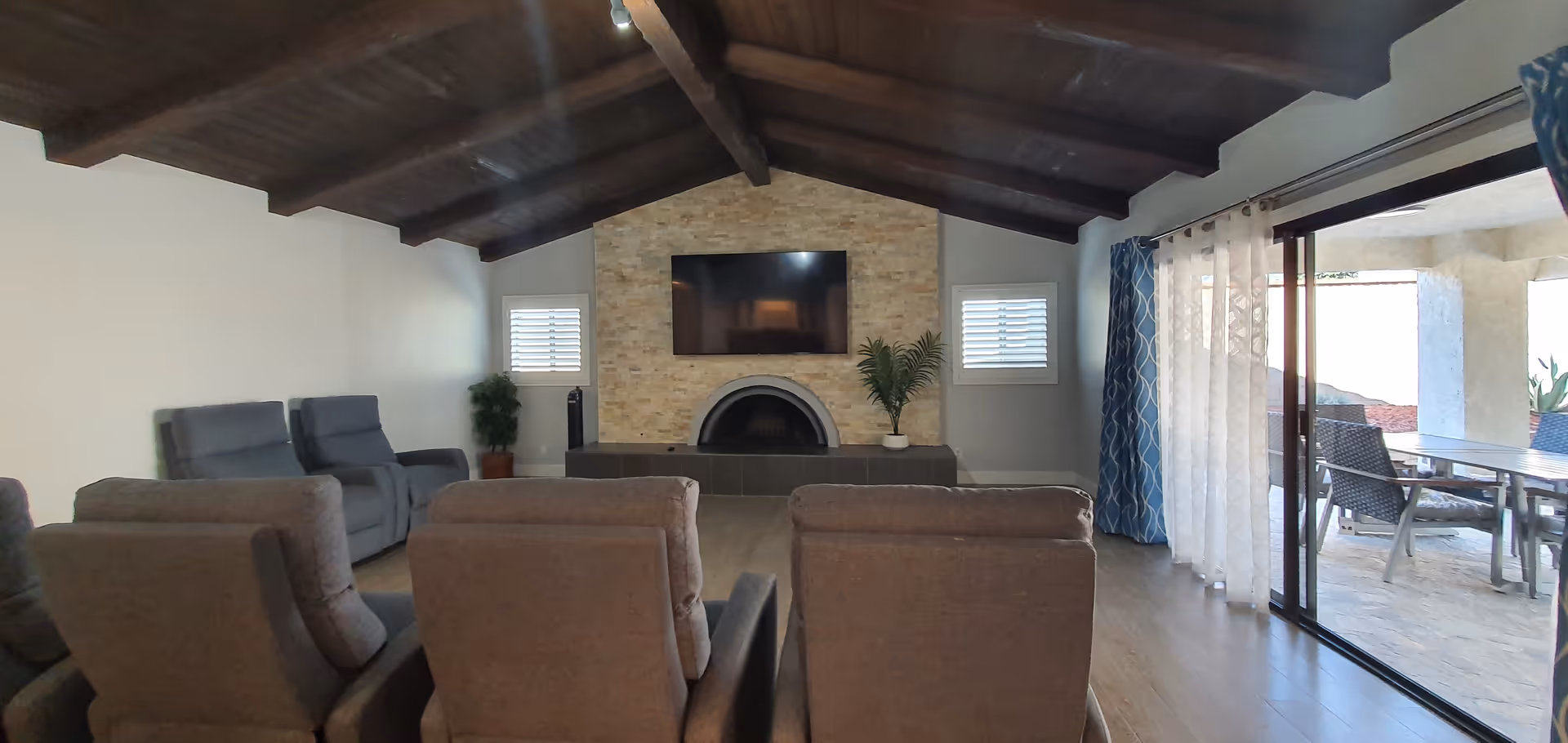 Interior view of a senior living facility common area with a vaulted wooden ceiling, a stone fireplace with a mounted flat-screen TV above it, two small windows on either side, several cushioned recliner chairs arranged facing the fireplace, and sliding glass doors leading to an outdoor patio with a table and chairs.