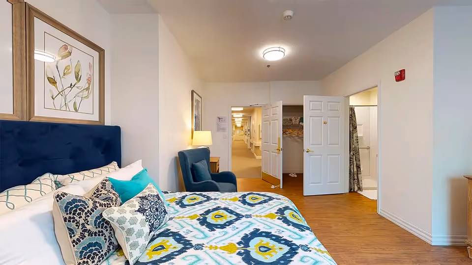 Well-lit bedroom with a patterned bed, an armchair, and open doors leading to a hallway and bathroom.