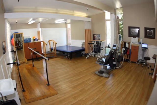 A rehabilitation room with wooden flooring featuring parallel bars for walking exercises, a blue therapy table, exercise equipment including a recumbent bike, and various medical and therapy supplies. The room has beige walls, large windows, and framed pictures on the wall.