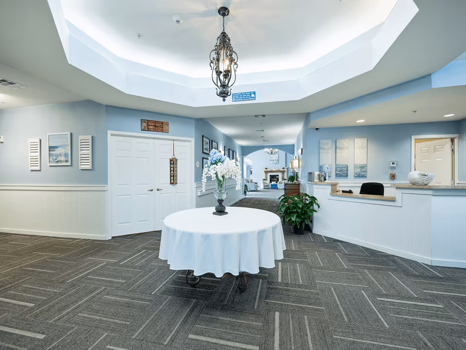 Interior view of a senior living facility lobby with a round table covered with a white cloth and a vase of flowers in the center. The walls are painted light blue with white wainscoting, and there is a chandelier hanging from a recessed ceiling. To the right is a reception desk with decorative items and a plant. In the background, there is a hallway leading to a seating area with a fireplace.