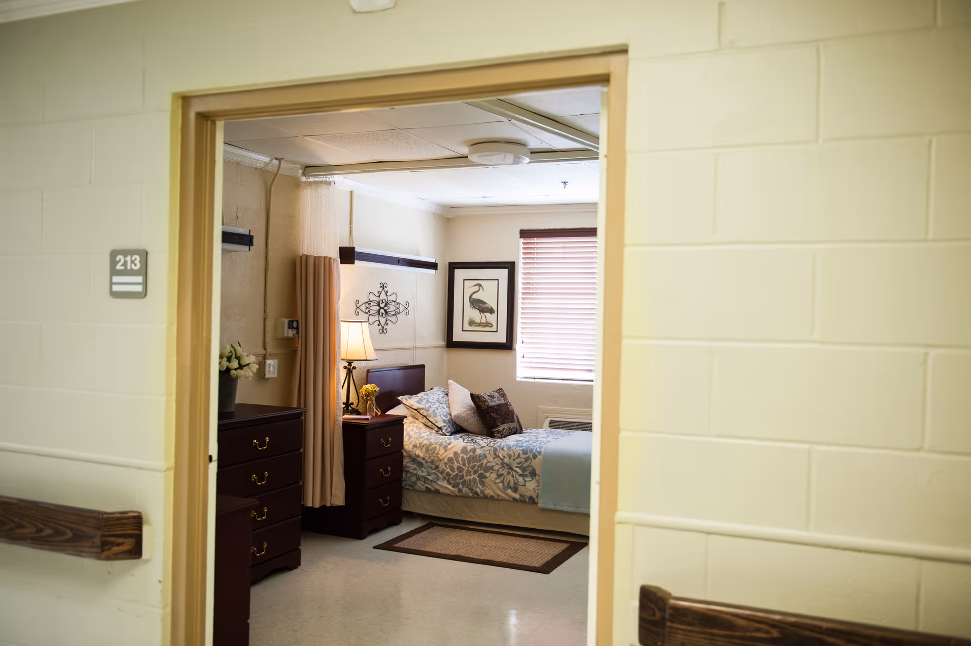 View through a doorway into a furnished bedroom with a bed, bedside tables and lamp, dressers, wall art, and a hallway sign reading 213.