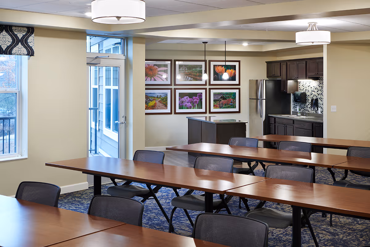Interior view of a communal dining or meeting room with multiple wooden tables and black mesh chairs. The room features a small kitchen area with dark cabinets, a stainless steel refrigerator, a sink, and a tiled backsplash. There are framed floral photographs on the wall and large windows letting in natural light.