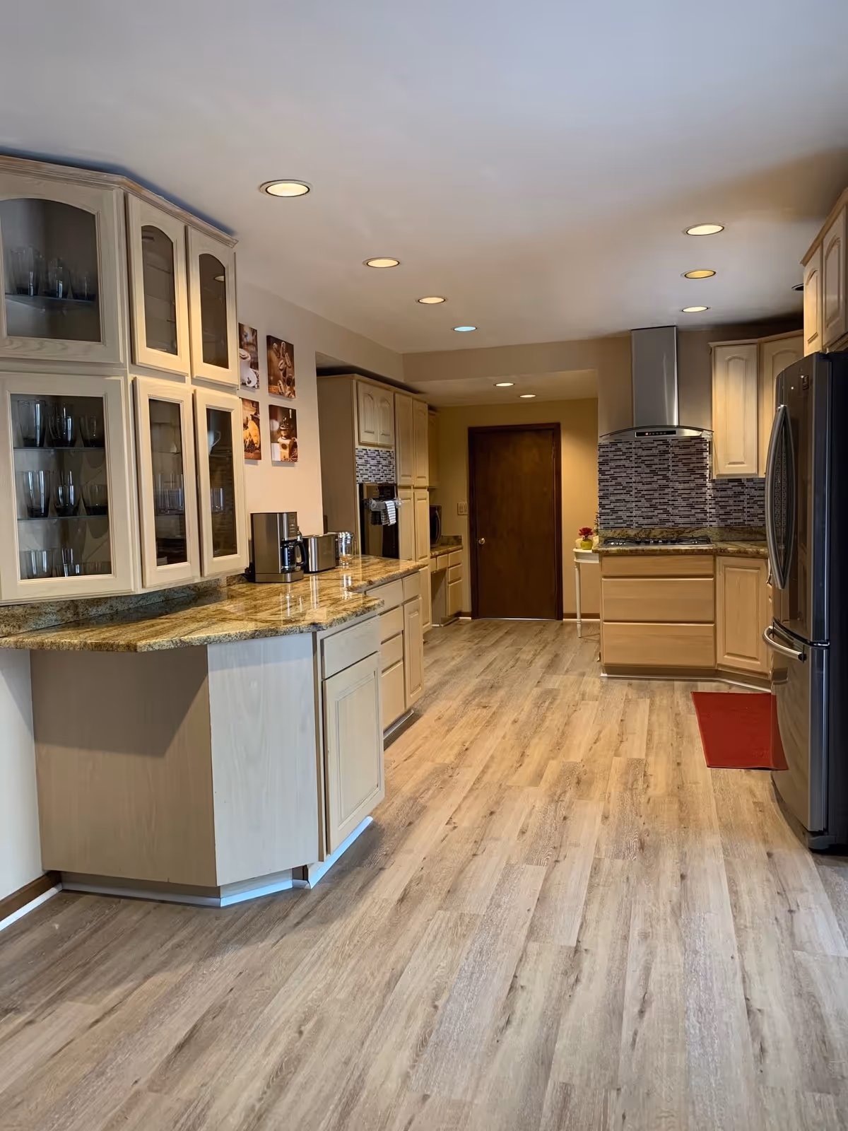 Open kitchen with light wood cabinetry, granite countertops, stainless steel refrigerator and range hood, and wood-look flooring.