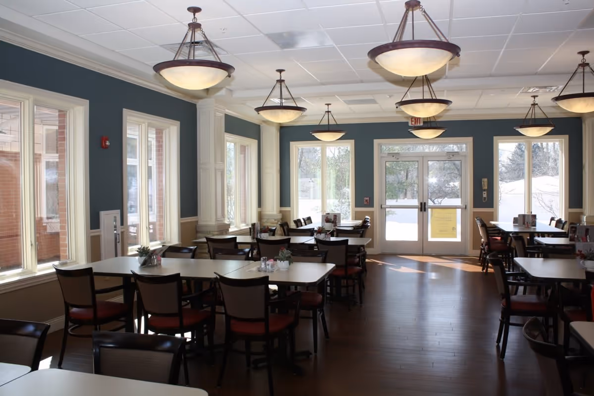 A bright dining room in a senior living facility with multiple tables and chairs arranged neatly. Large windows allow natural light to fill the room, and several hanging light fixtures provide additional illumination. The walls are painted blue with white trim, and the floor is wooden. The entrance door is visible at the far end of the room.