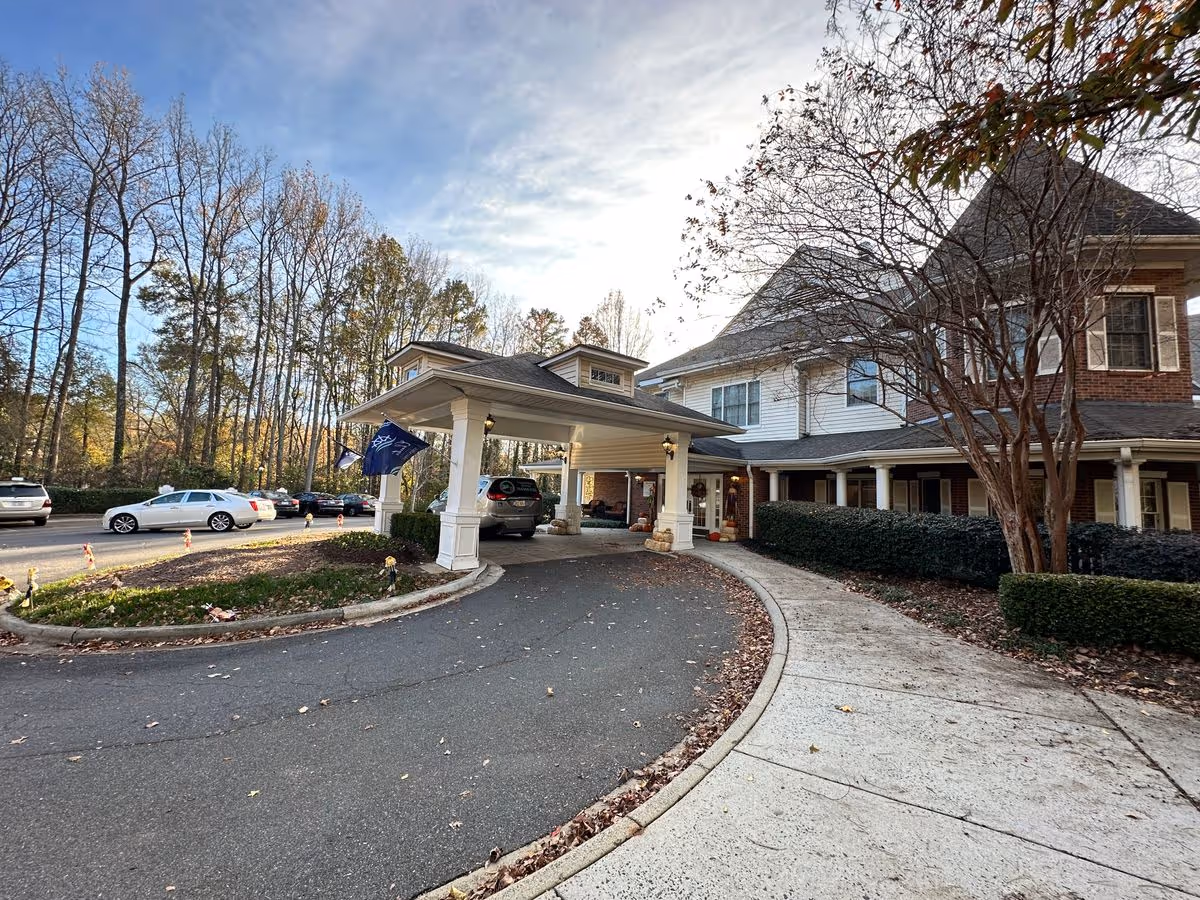 Front entrance of a senior living building featuring a covered porte-cochere, parked cars, and trees lining the driveway.