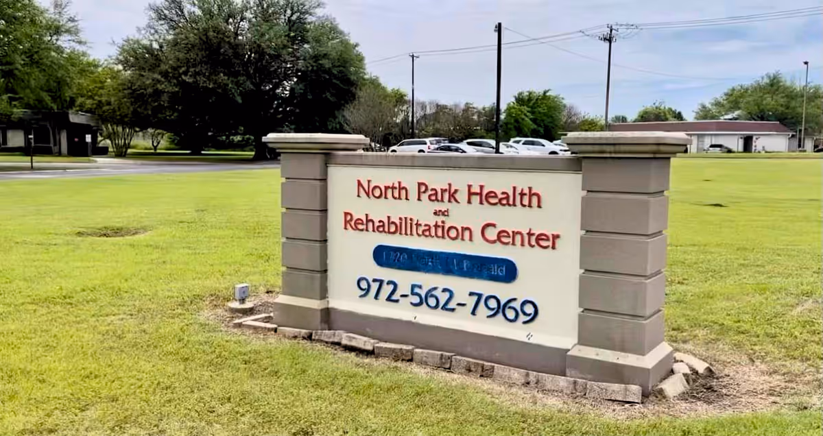 Outdoor view of a large sign for North Park Health and Rehabilitation Center on a grassy area with trees, parked cars, and buildings in the background.