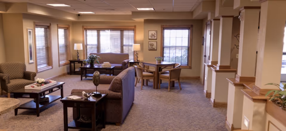 A cozy living room area in a retirement community with multiple seating options including armchairs and sofas, wooden tables with decorative items, large windows with blinds allowing natural light, and warm beige walls and carpet.
