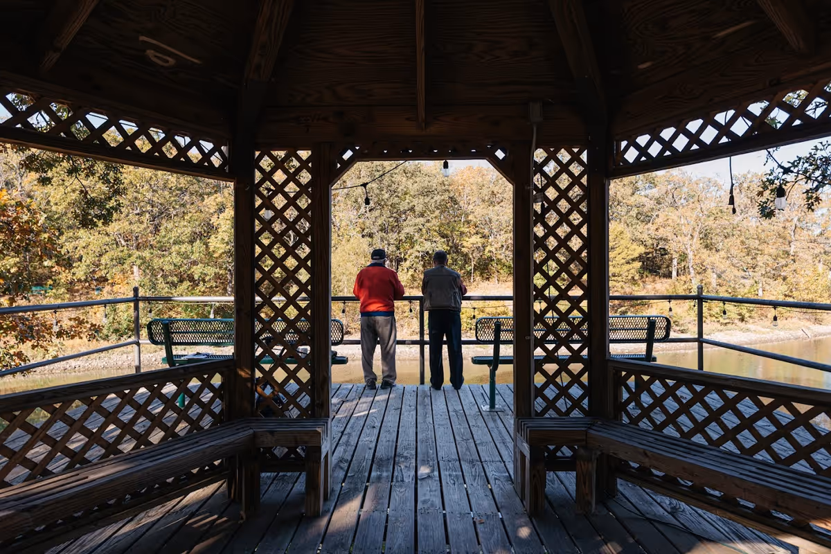 Two elderly men standing on a wooden gazebo deck overlooking a pond surrounded by trees with autumn foliage. The gazebo has lattice woodwork and benches along the sides.