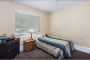 A small bedroom with a single bed covered in a striped comforter, a wooden nightstand with a lamp, a dark wooden dresser with decorative items, and a window with closed blinds.