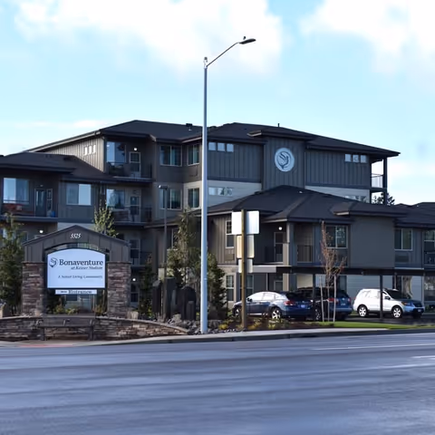 Exterior view of Bonaventure at Keizer Station, a senior living community building with multiple floors, balconies, and parked cars in front. A stone sign with the facility name is visible near the street.