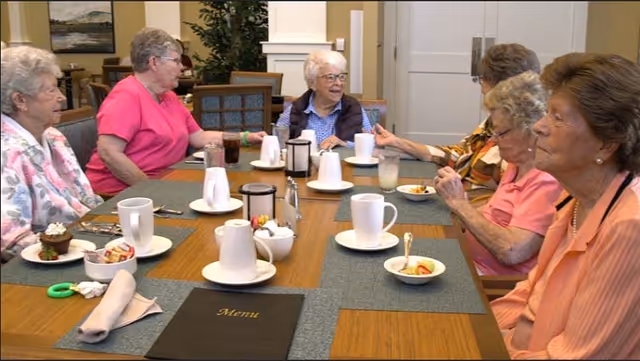 A group of elderly women sitting around a dining table in a senior living facility, enjoying conversation and light refreshments including coffee and small bowls of fruit. The table is set with cups, plates, and a menu, and the room has a warm, inviting atmosphere.
