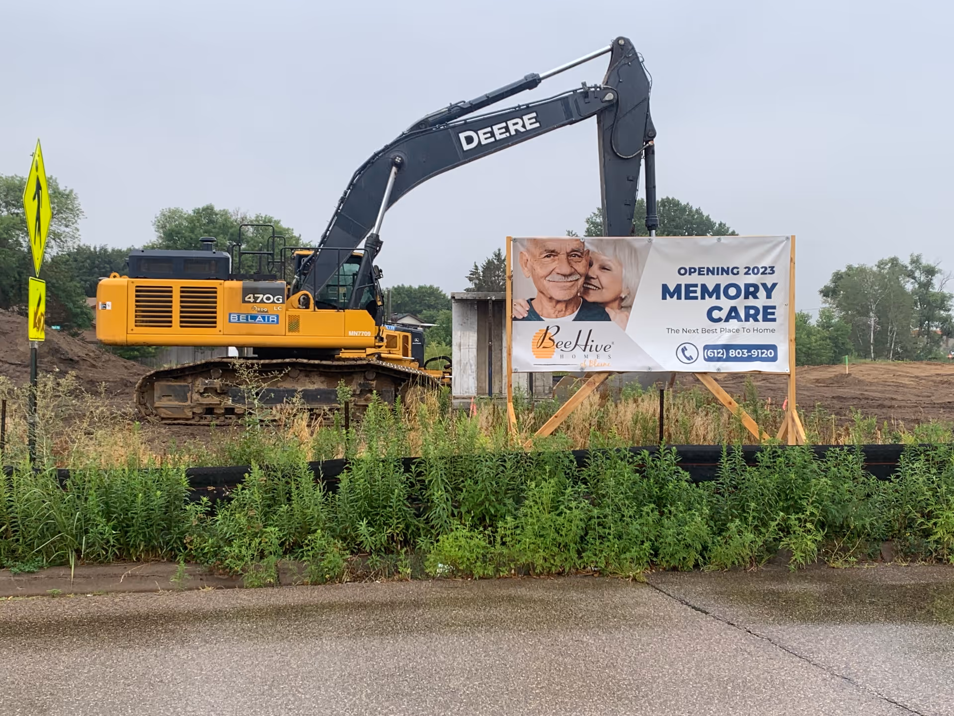 A construction site with a large yellow and black Deere excavator parked next to a sign announcing the opening of a memory care facility in 2023. The sign features an elderly couple smiling and the text 'BeeHive Homes, Opening 2023 Memory Care, The Next Best Place To Home' along with a phone number. There is greenery and a pedestrian crossing sign nearby.