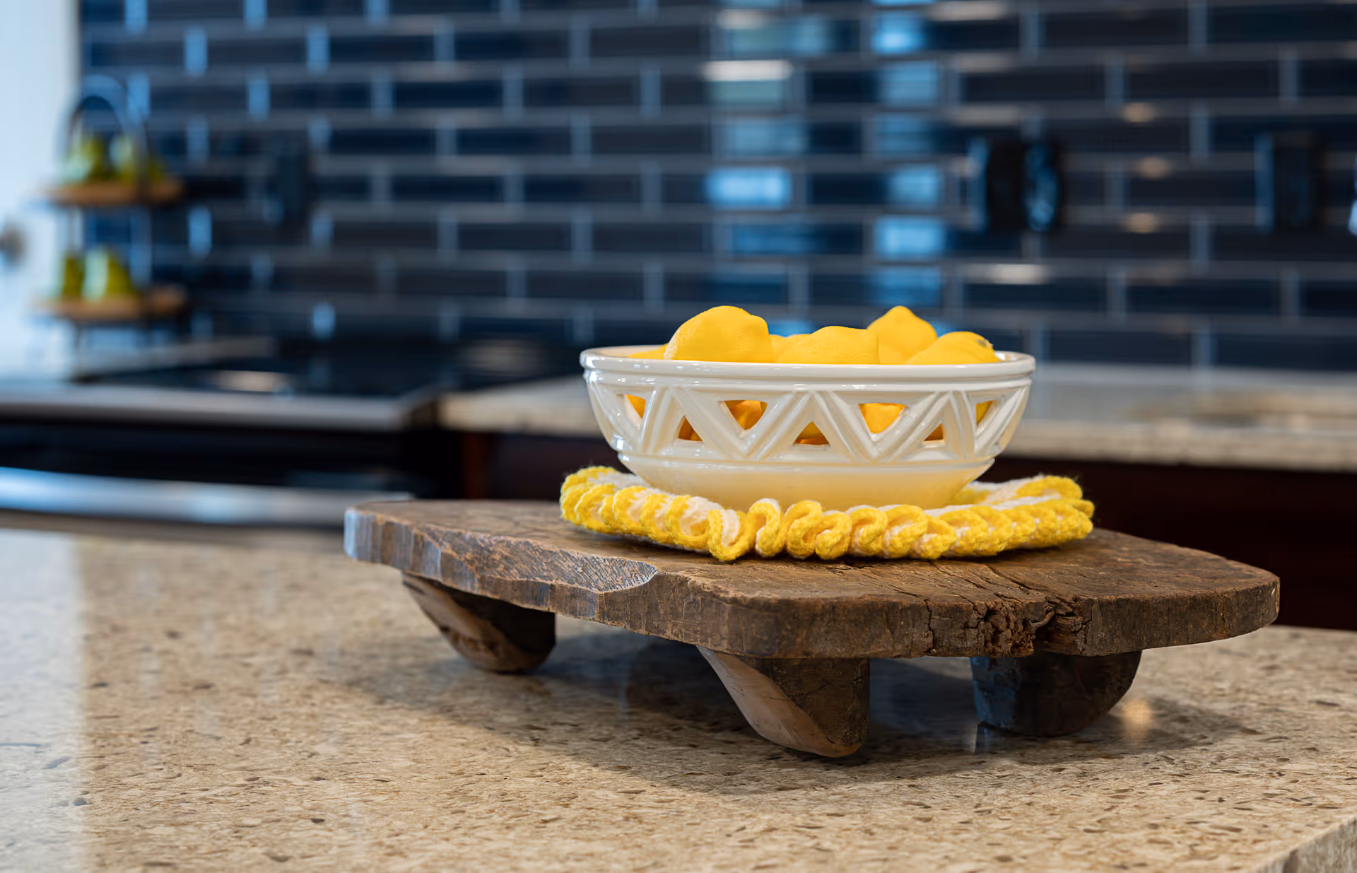A kitchen countertop with a rustic wooden stand holding a white bowl of lemons on a yellow trivet, with a tiled backsplash and stovetop in the background.