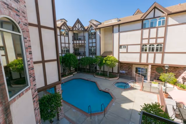 View of an outdoor courtyard at Lexington Assisted Living featuring a swimming pool, a hot tub, surrounding trees, and multi-story building with Tudor-style architecture under a clear blue sky.