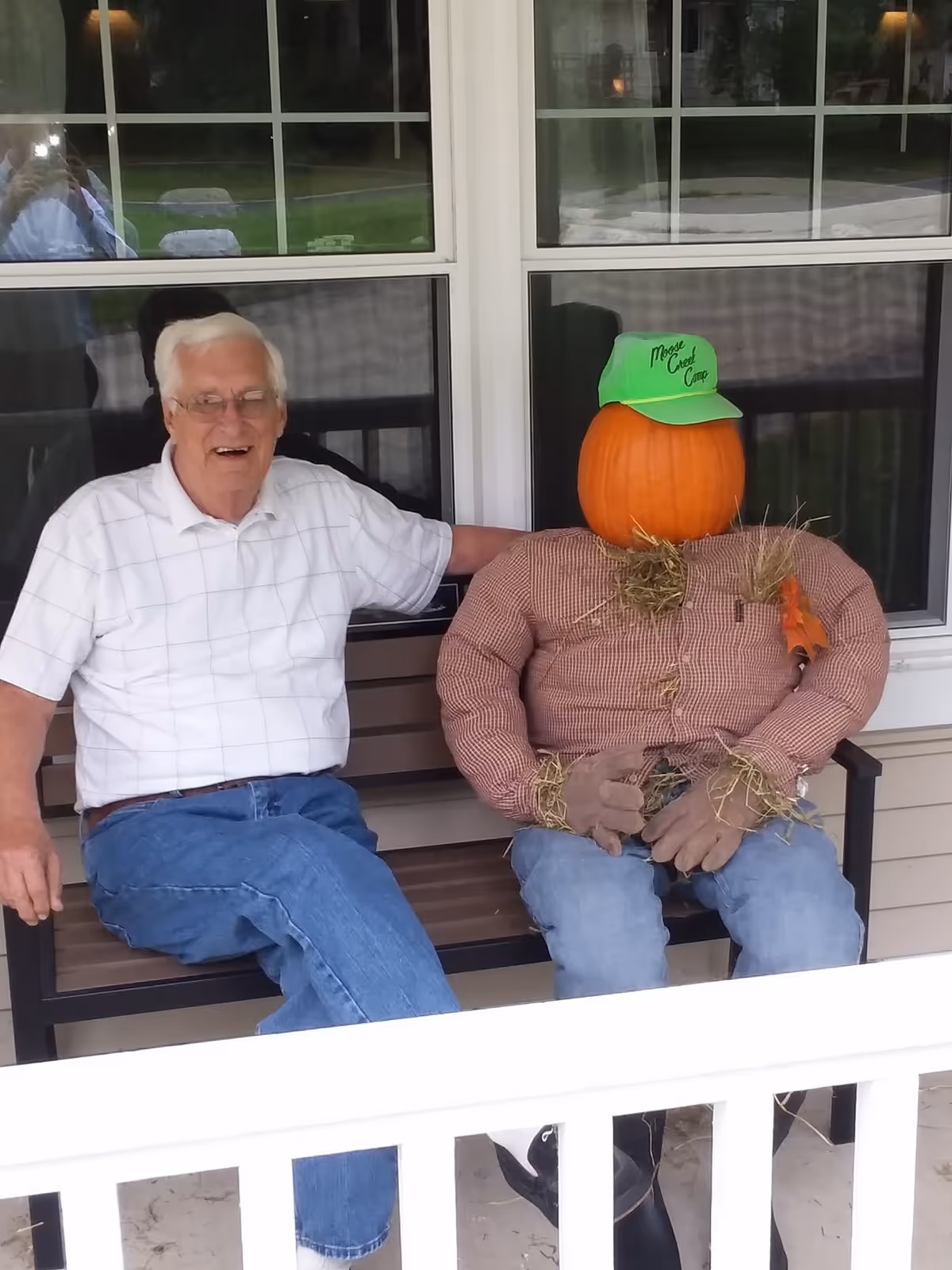 An elderly man sits on a porch bench next to a scarecrow with a pumpkin head wearing a green cap.