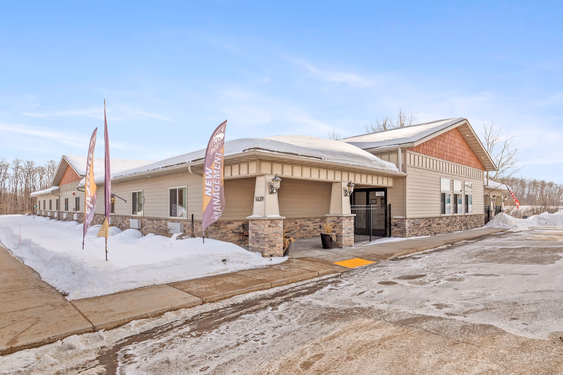 Exterior front of a single-story senior living facility with stone accents, flags in the snow, and a covered entrance.