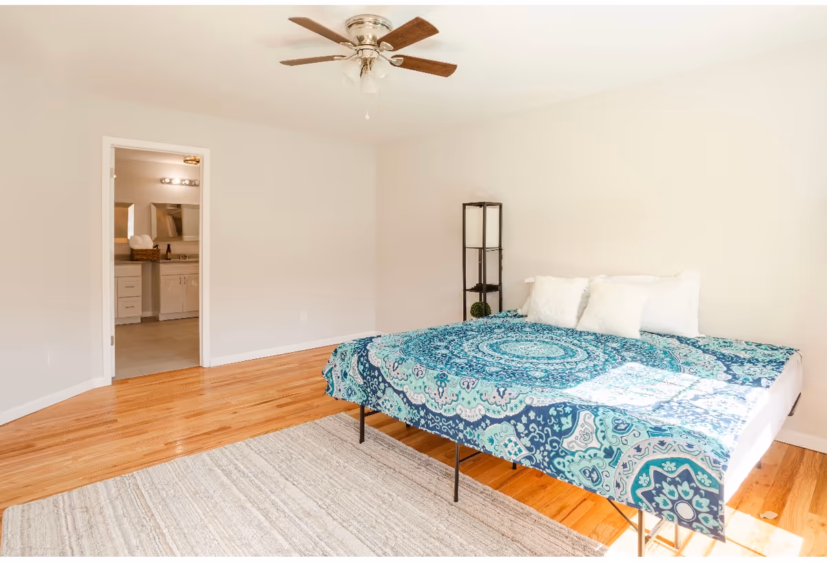 A bright bedroom with a wooden floor, a bed covered with a blue and turquoise patterned bedspread, three white pillows, a beige rug, a ceiling fan with wooden blades, and an open door leading to a bathroom with a vanity and mirror.