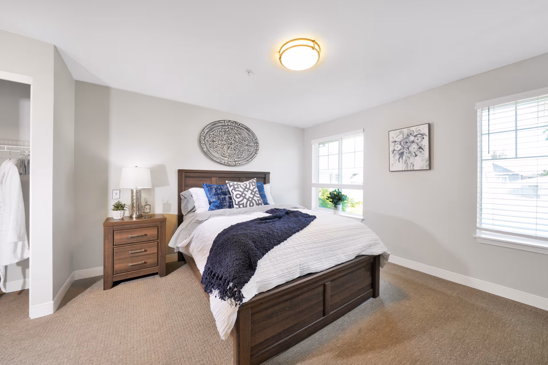 A bright and clean bedroom with a wooden bed frame and a neatly made bed featuring white and blue bedding. There is a wooden nightstand with a lamp, a small plant, and a clock. The room has beige carpet, light gray walls, two windows with white blinds, a decorative wall piece above the bed, and a closet with white robes hanging inside.