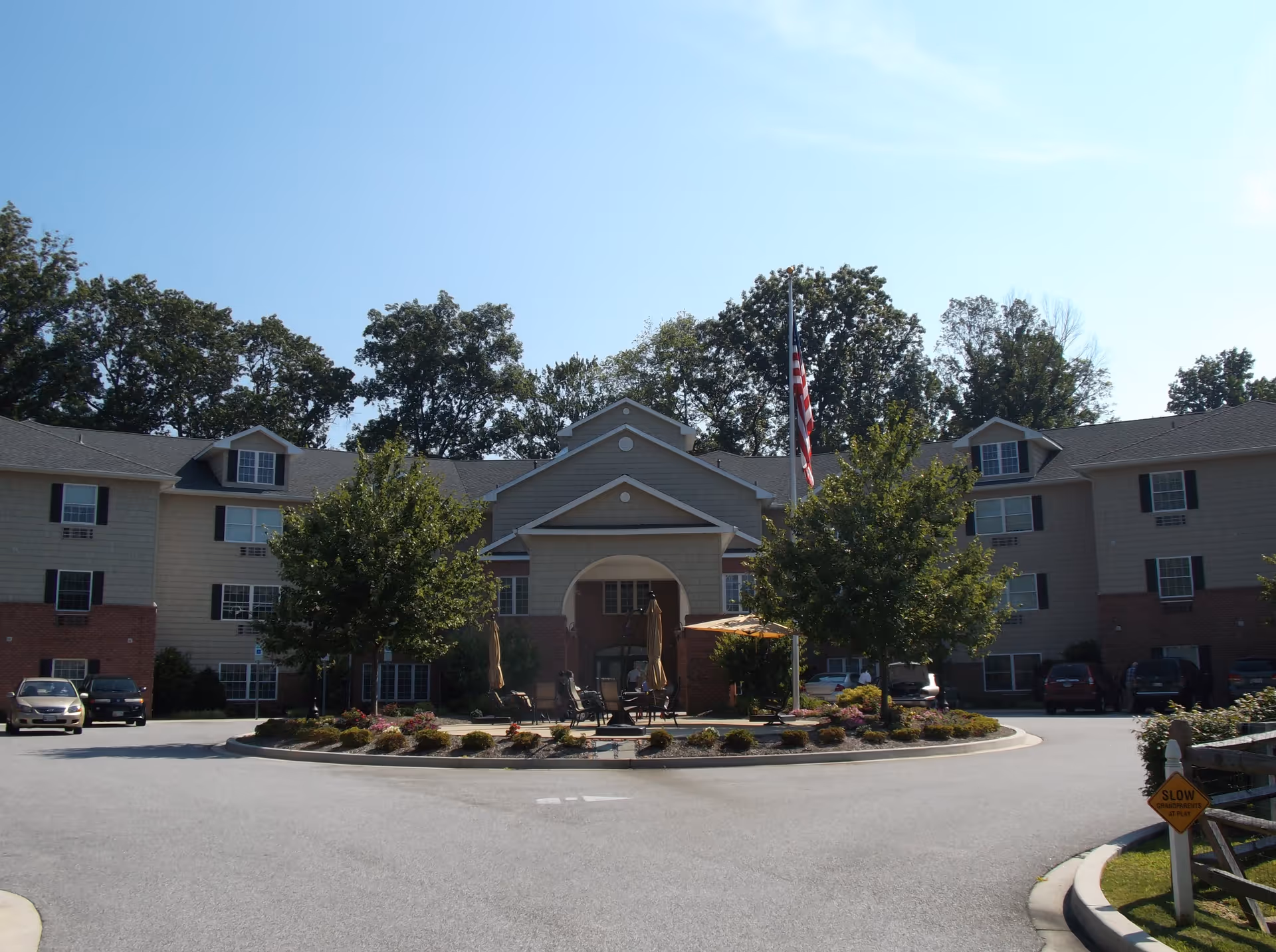 Front exterior view of a multi-story senior living facility building with a circular driveway, landscaped center island with trees and flowers, outdoor seating with umbrellas, several parked cars, and an American flag on a flagpole.