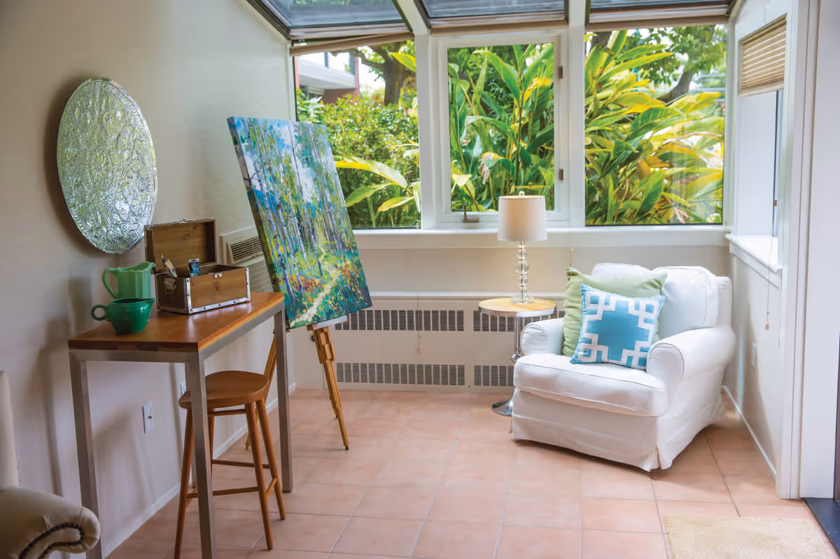 Bright sunroom-style living area with a white armchair, side table and lamp, a small desk and stool, and an easel with a painting in front of large windows showing lush greenery.