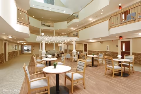 A spacious, well-lit common area in a senior living facility with multiple round tables and wooden chairs arranged neatly. The room features a high ceiling with a skylight, a decorative white pergola structure in the center, and a mix of carpet and wood flooring. The walls are light-colored with wooden paneling halfway up, and there are doors and railings visible on the upper floors.