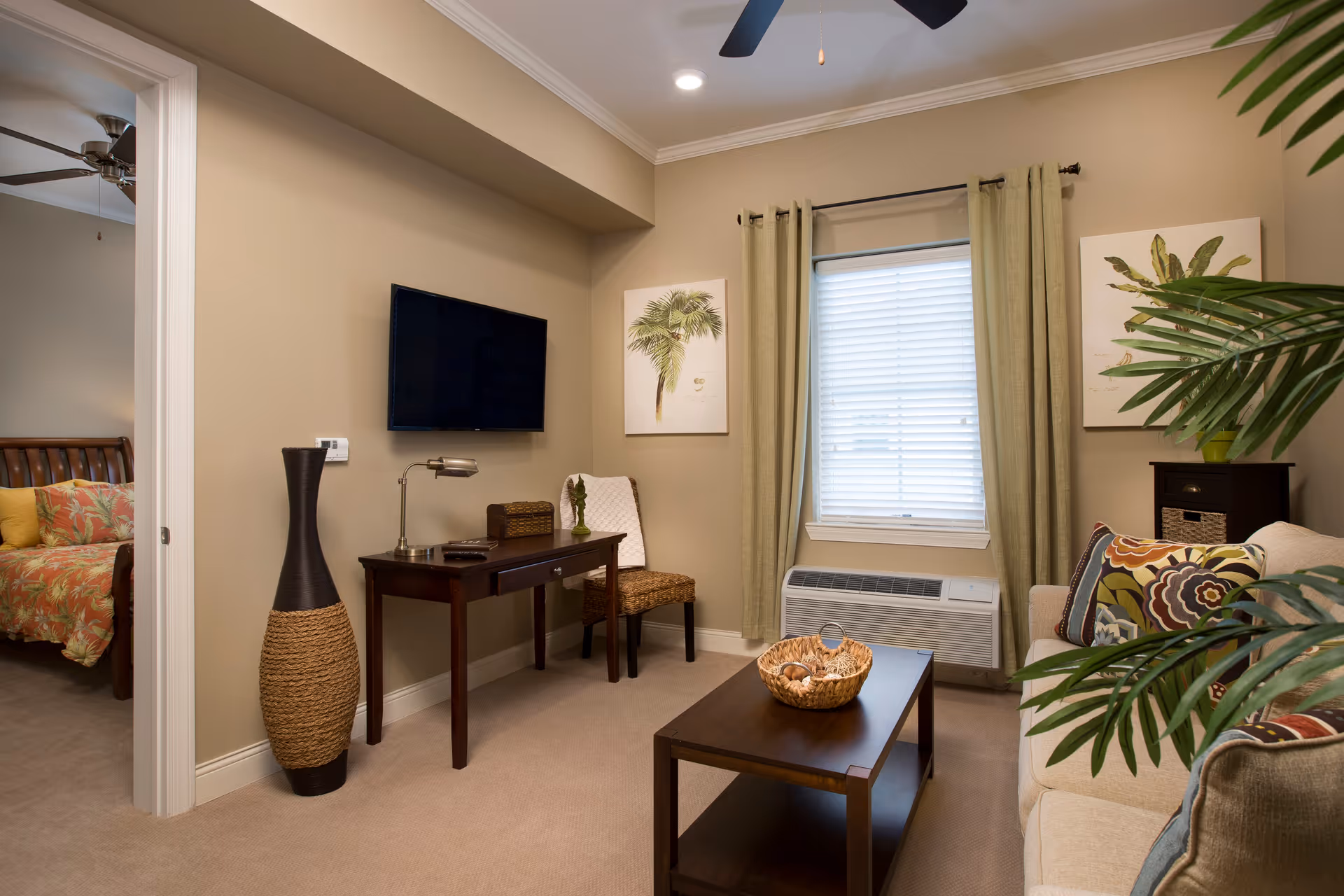 A cozy living room with beige walls and carpet, featuring a beige sofa with colorful patterned cushions, a dark wooden coffee table with a decorative basket, a window with green curtains and blinds, a wall-mounted flat-screen TV above a dark wooden desk with a chair, a tall decorative vase, and framed botanical artwork on the walls. A bedroom with a bed covered in a floral bedspread is visible through an open doorway.