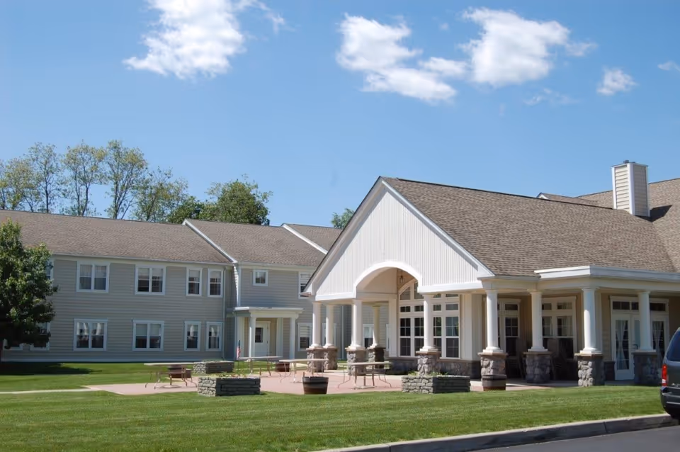 Front exterior of an assisted living building with a covered columned entrance, patio seating, and a manicured lawn under a blue sky.