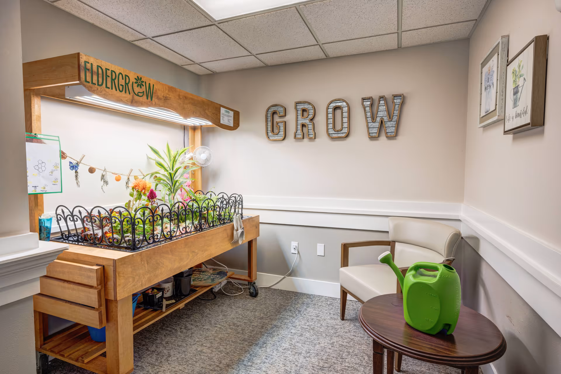 Indoor gardening station with plants growing under a wooden frame labeled 'ELDERGROW'. A small fan is attached to the frame. On the wall behind, large decorative letters spell out 'GROW'. A beige armchair is next to a small round wooden table with a green watering can on it. The room has light-colored walls and carpeted floor.