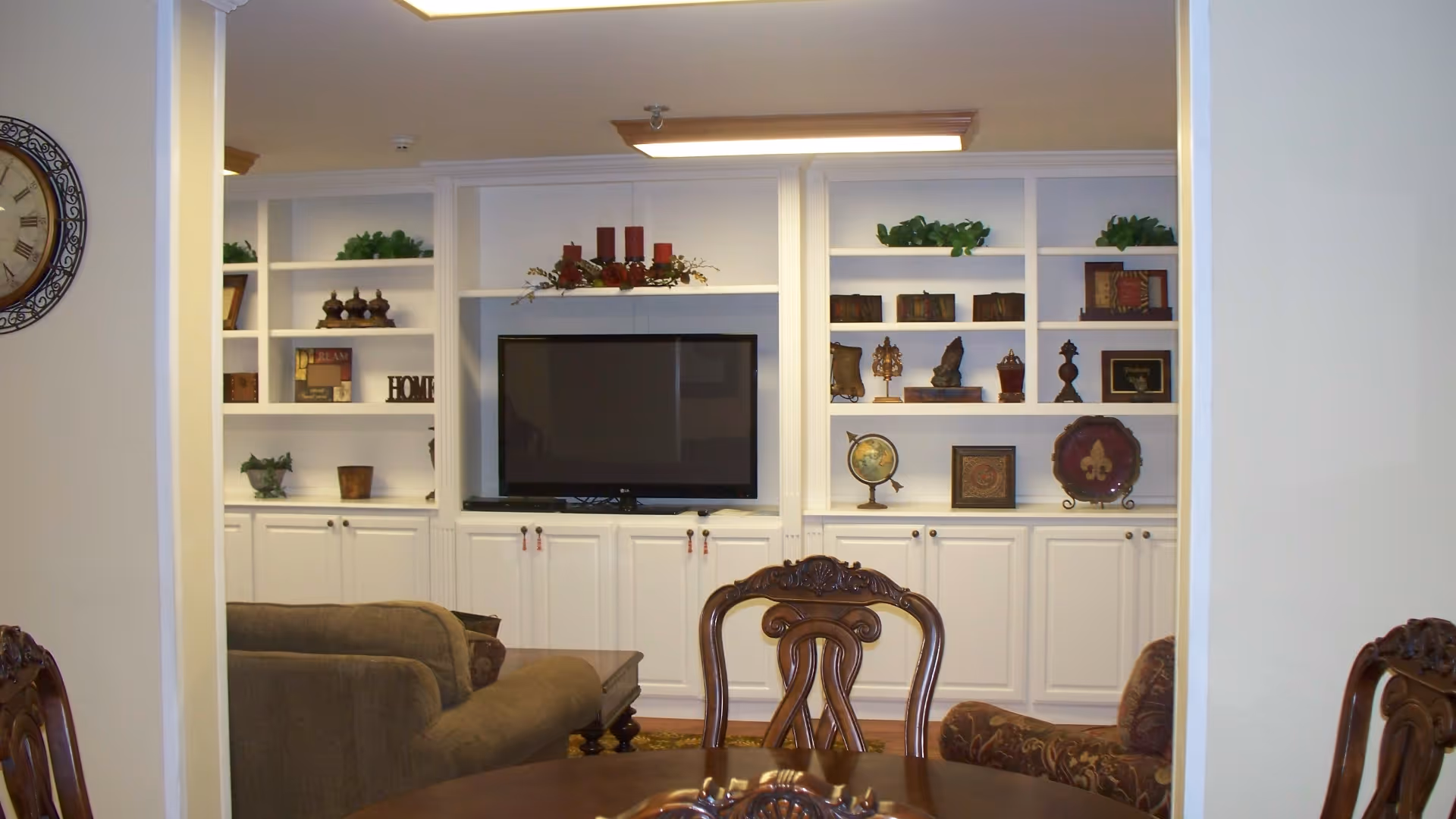A cozy living room area with a large flat-screen TV mounted in a white built-in shelving unit filled with decorative items and plants. In front of the TV, there are upholstered armchairs and a wooden dining table with carved wooden chairs. A large wall clock is partially visible on the left side.