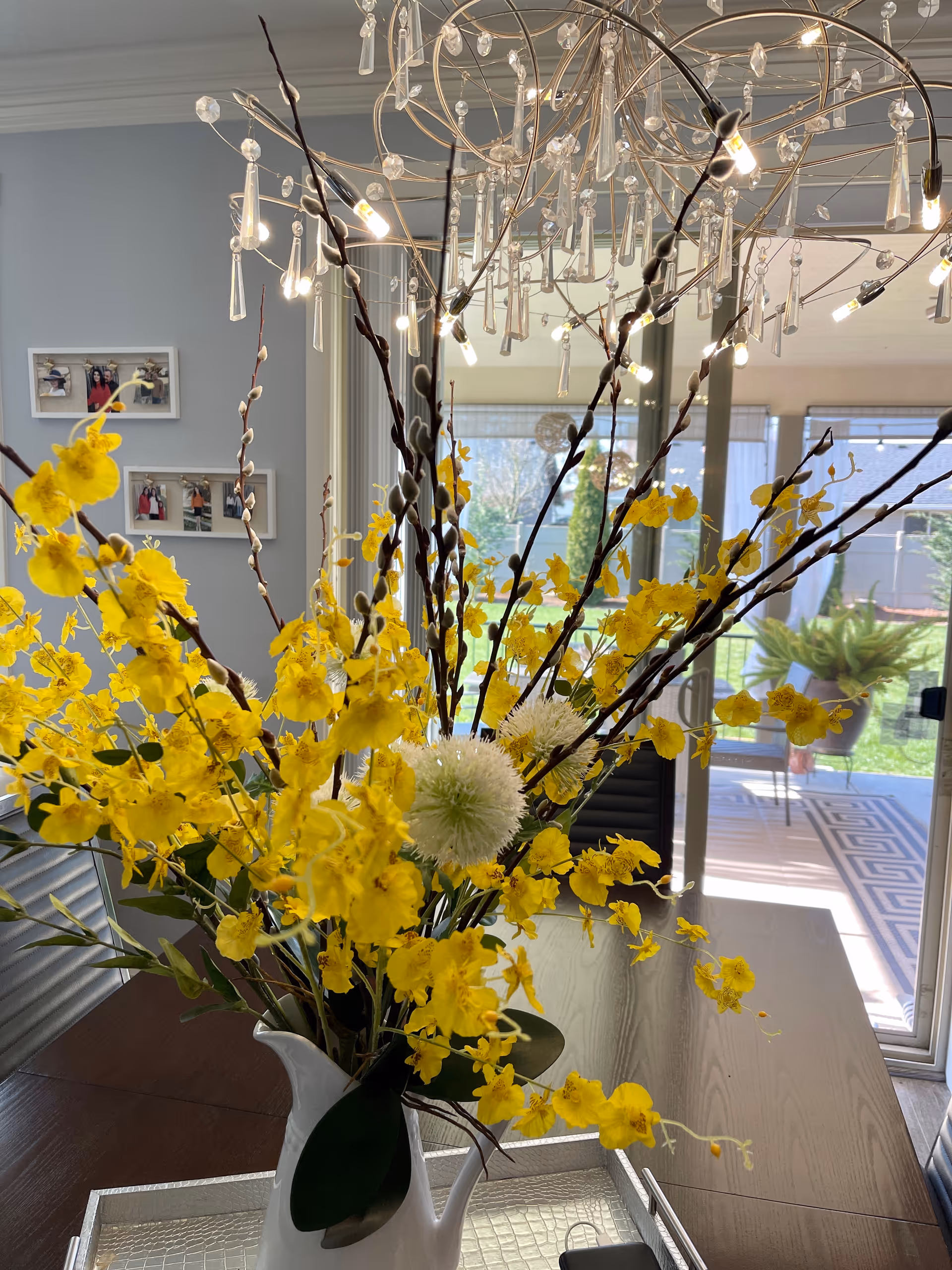 A close-up view of a floral arrangement with bright yellow flowers and white spherical blooms in a white pitcher vase on a dining table. Behind the flowers, a modern chandelier with hanging crystal decorations is visible. In the background, there is a glass sliding door leading to an outdoor patio with greenery and a patterned outdoor rug. On the left wall, there are framed photos.