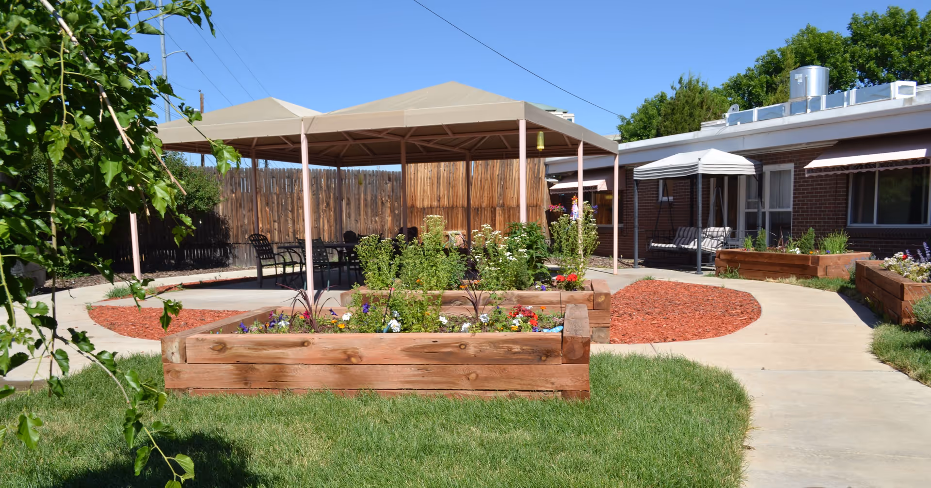 Outdoor garden area with raised wooden flower beds filled with plants and flowers, a paved walkway, a shaded seating area with chairs under a canopy, and a brick building with windows and awnings in the background.