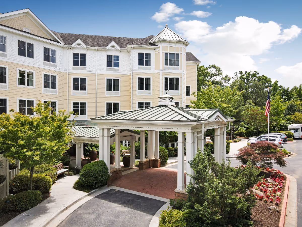 Exterior view of a senior living facility with a covered entrance, surrounded by landscaped greenery, trees, and flower beds. Several parked cars and an American flag are visible in the parking area under a partly cloudy sky.