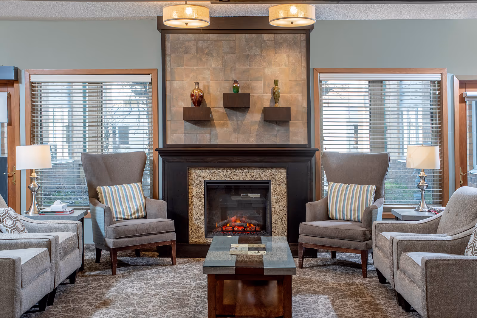 A cozy senior living facility common area with a central fireplace surrounded by four gray upholstered armchairs, each with a decorative pillow. Two side tables with lamps flank the chairs, and large windows with blinds allow natural light to enter the room. The fireplace mantle features three decorative vases on small shelves, and a patterned carpet covers the floor.