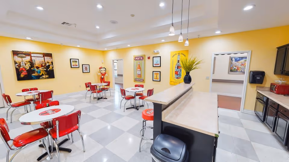 A bright dining area with yellow walls and checkered gray and white floor tiles. The room features several round tables with red chairs and stools, some tables have Coca-Cola logos on them. There is a counter with a beige countertop and a black base, with a vase containing green plants on it. The walls are decorated with vintage-style posters and artwork, including a large Betty Boop figure near the back wall. The room also includes a kitchenette area with dark cabinets, a red microwave, and a black paper towel dispenser.