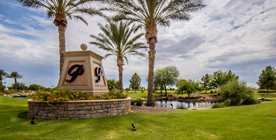 A landscaped outdoor area with green grass, palm trees, and a small pond. In the foreground, there is a stone planter with yellow flowers and a beige monument featuring the letter 'P' on multiple sides. The sky is partly cloudy.