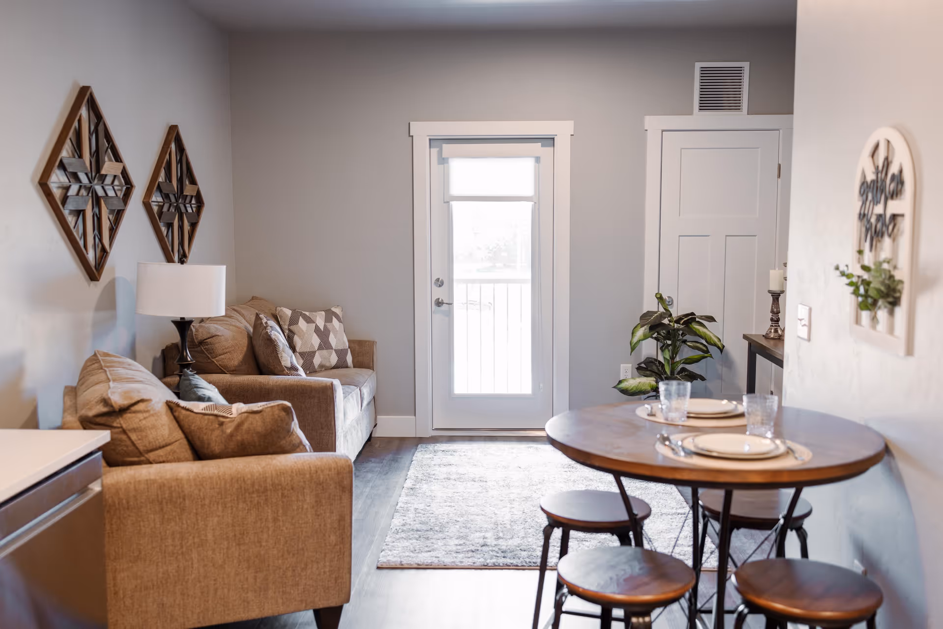 A cozy living area with two beige sofas adorned with patterned cushions, a floor lamp, and two decorative wooden wall hangings. A round wooden dining table with four stools is set with plates and glasses. A door with a window and a small rug are visible in the background, along with a potted plant and a small console table with candles.